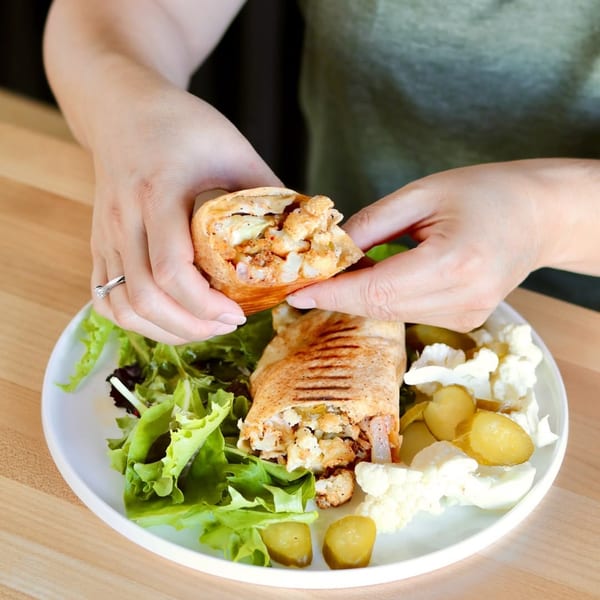 Hands holding a Wahib's Mediterranean Express chicken shawarma wrap, with salad and pickles on a plate.