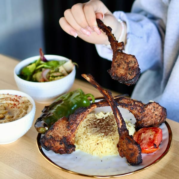 Hand holding a grilled lamb chop over a plate of rice, roasted vegetables, and hummus.